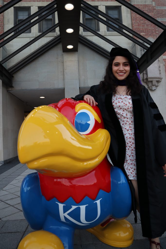 Samantha Natera in law graduate regalia next to a Kansas Jayhawk statue
